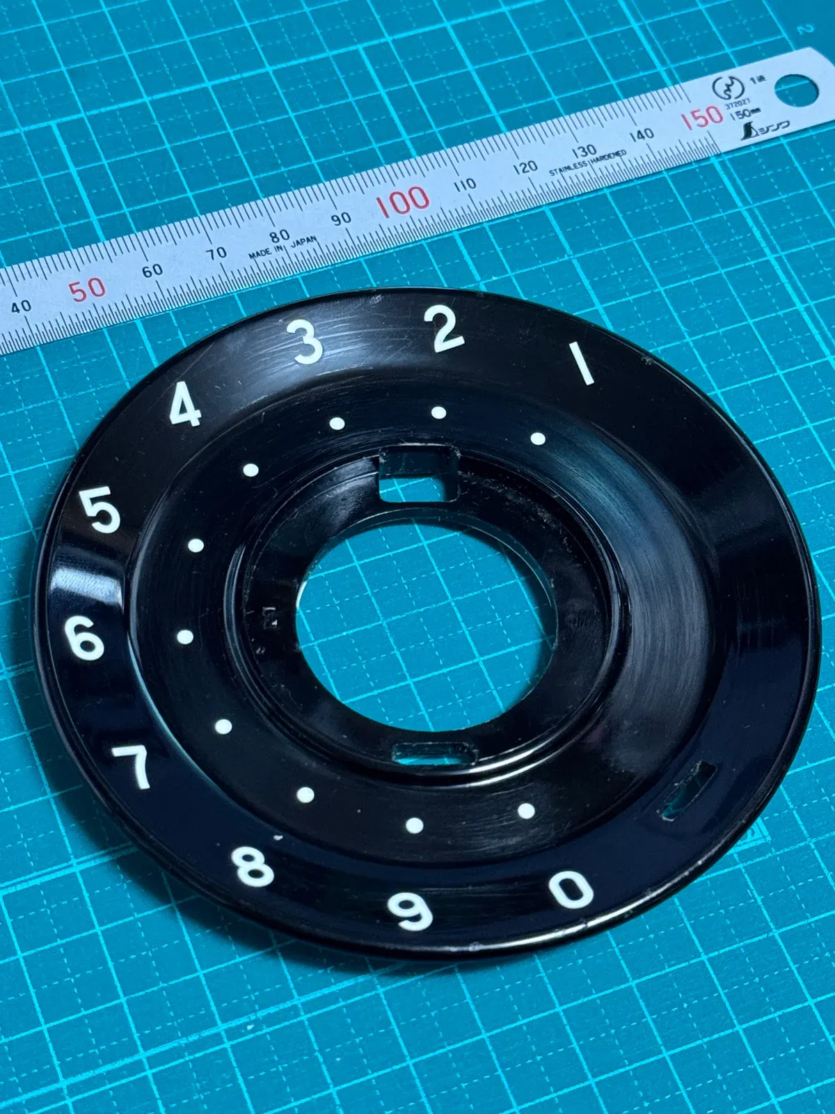 黒電話のダイヤル盤を定規と並べて撮った写真。黒い盤の外周に白い数字1〜9と0、内側に白い点が並ぶ