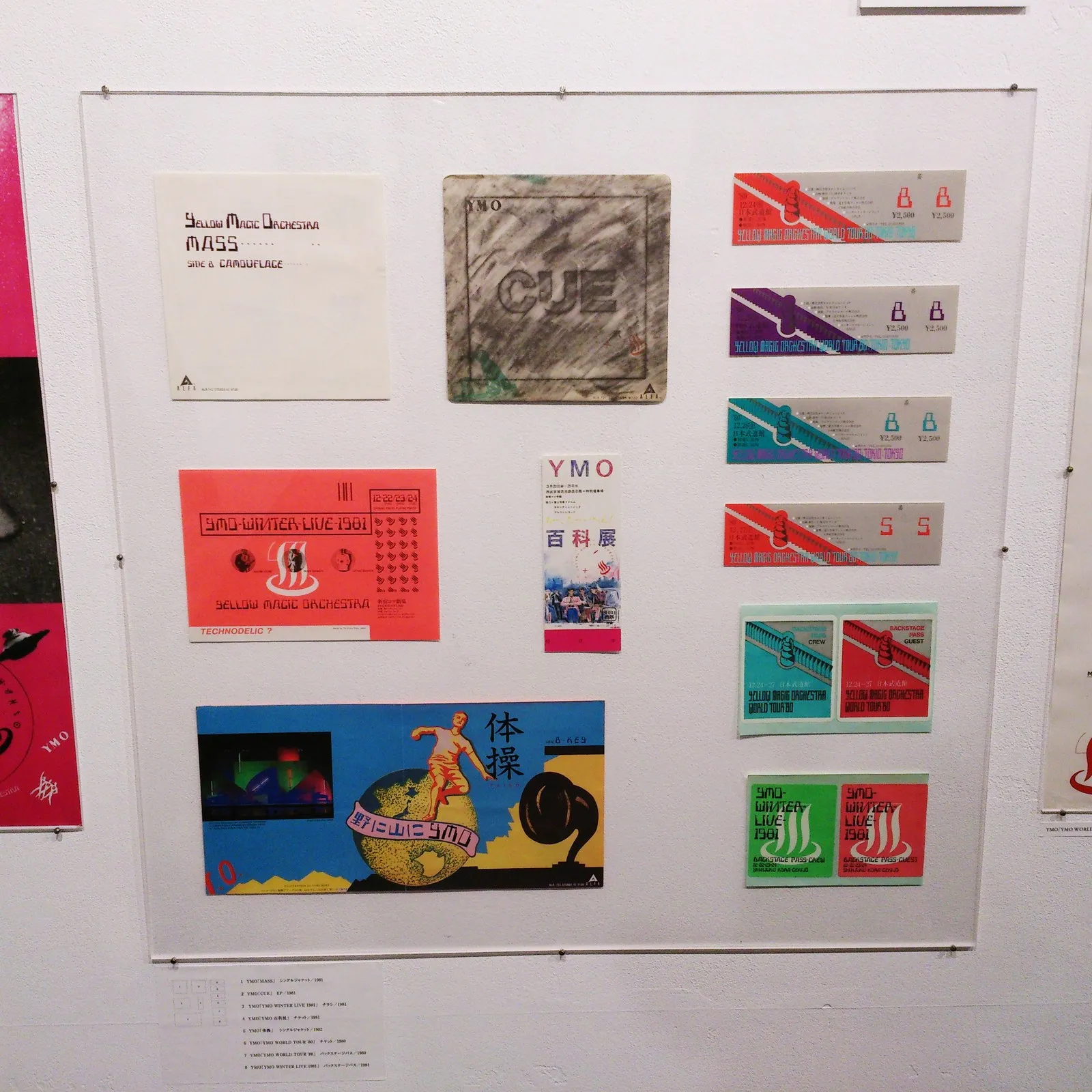 YMO の印刷物の展示。左上にシングル「MASS」のジャケット、中央に EP「CUE」、左に「YMO WINTER LIVE 1981」のチラシ、「YMO 百科展」のチケット、シングル「体操」のジャケット。右に1980年12月の日本武道館公演のチケット4枚(席種「B」「S」)と、バックステージパス2組